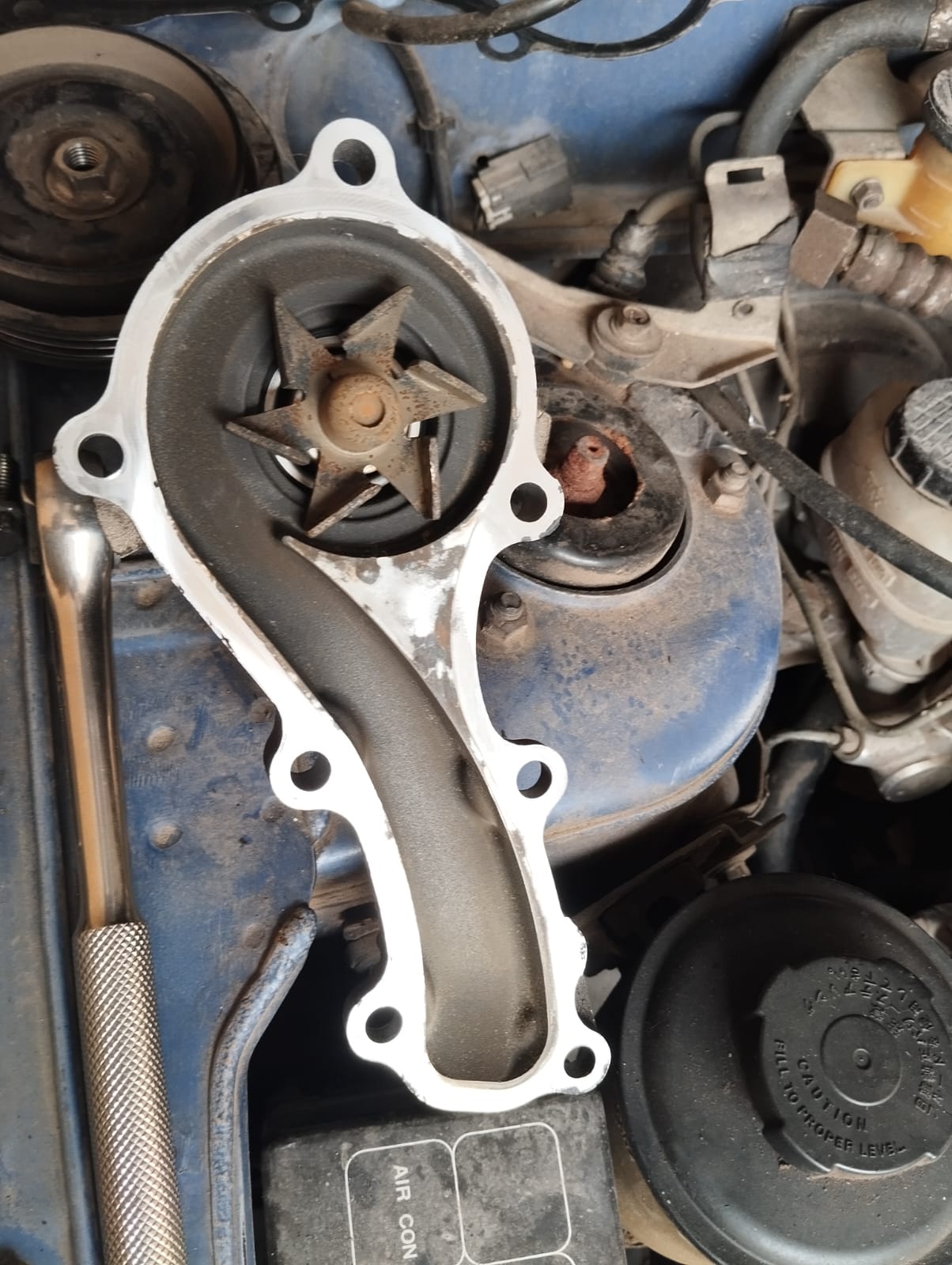 The water pump removed from the QG15 engine, its impeller vanes intact and still firmly fixed to the pulley shaft, held over the engine bay.